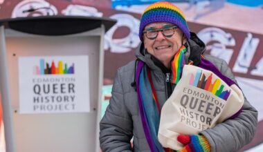 A man wearing a rainbow toque and mitts holds a back that reads 'Edmonton Queer History Project'
