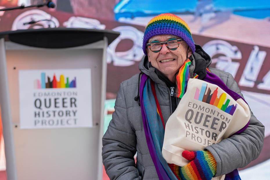 A man wearing a rainbow toque and mitts holds a back that reads 'Edmonton Queer History Project'