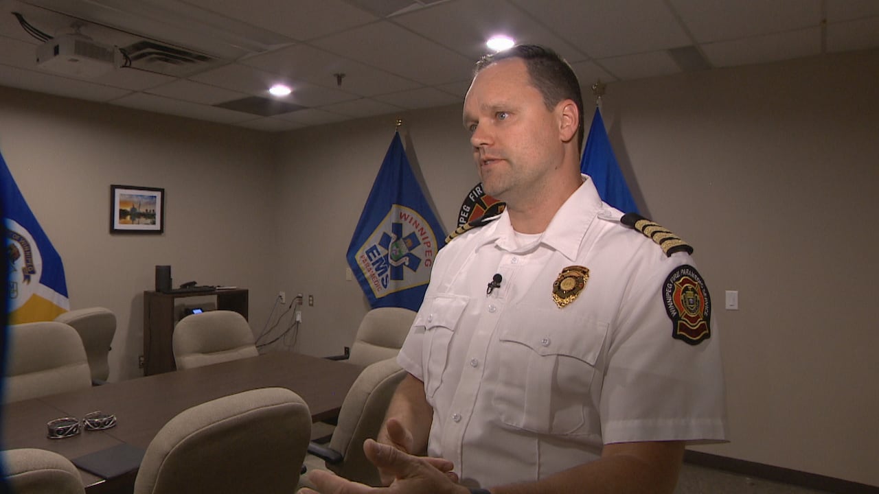A man wearing a white fire uniform with a badge and shoulder patches is pictured talking in a bedroom standing in front of 3 flags and a fire logo.