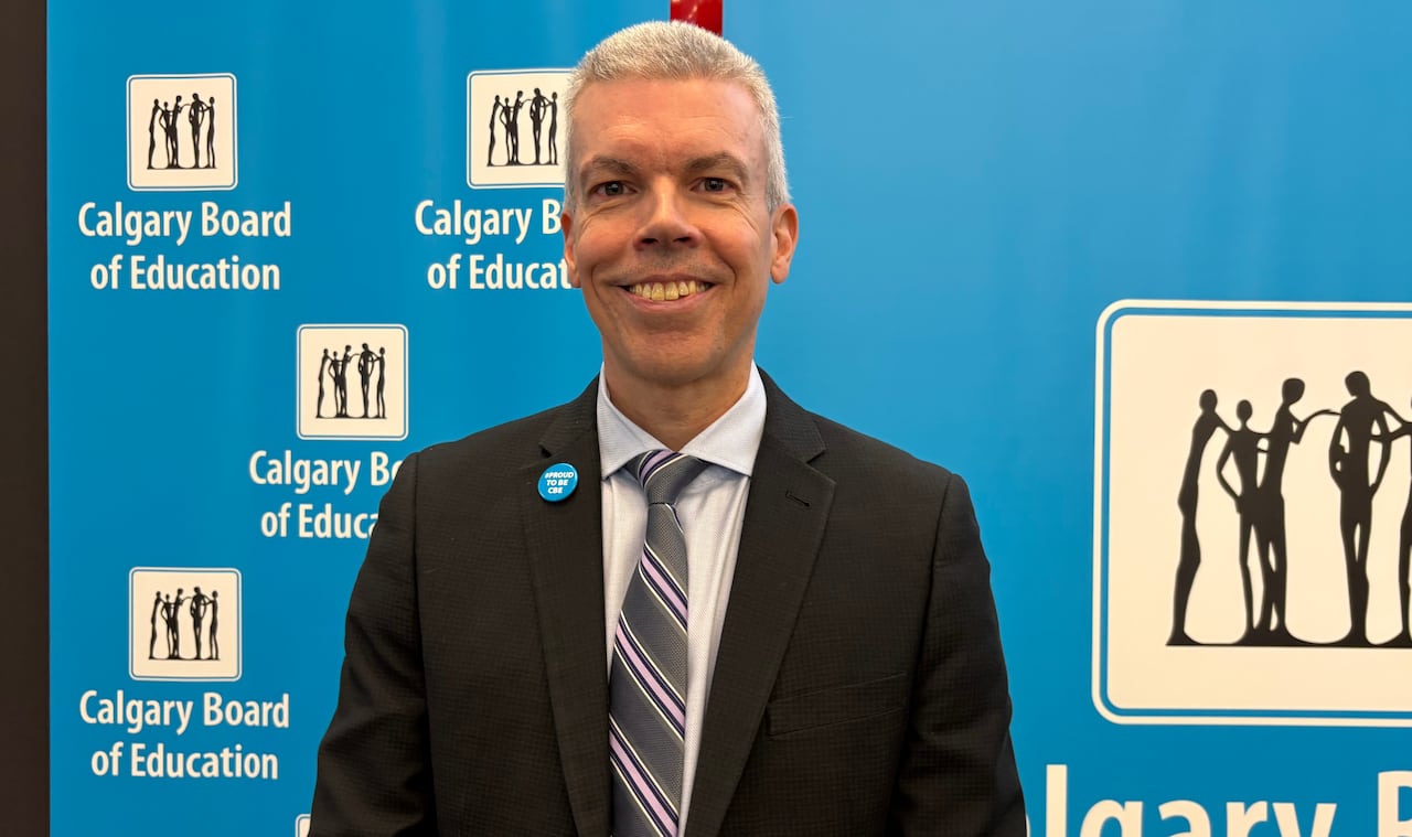A man wearing a suit smiles at the camera while standing in front of bright blue signs that say Calgary Board of Education