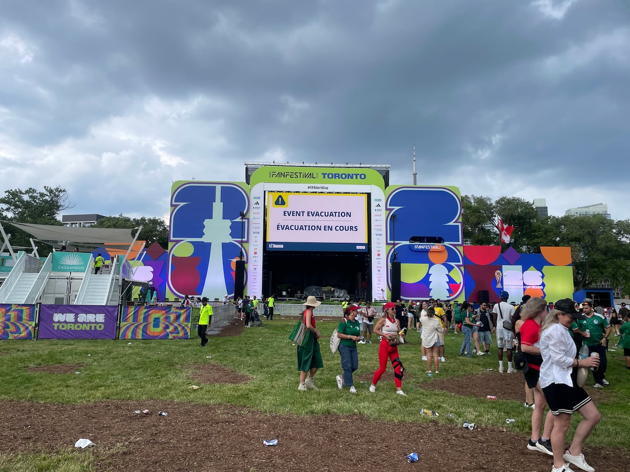 On a grey day threatening rain, a sparse crowd of people walk away from the FIFA Fan Festival Stage on a grassy field in Fort York in Toronto.
