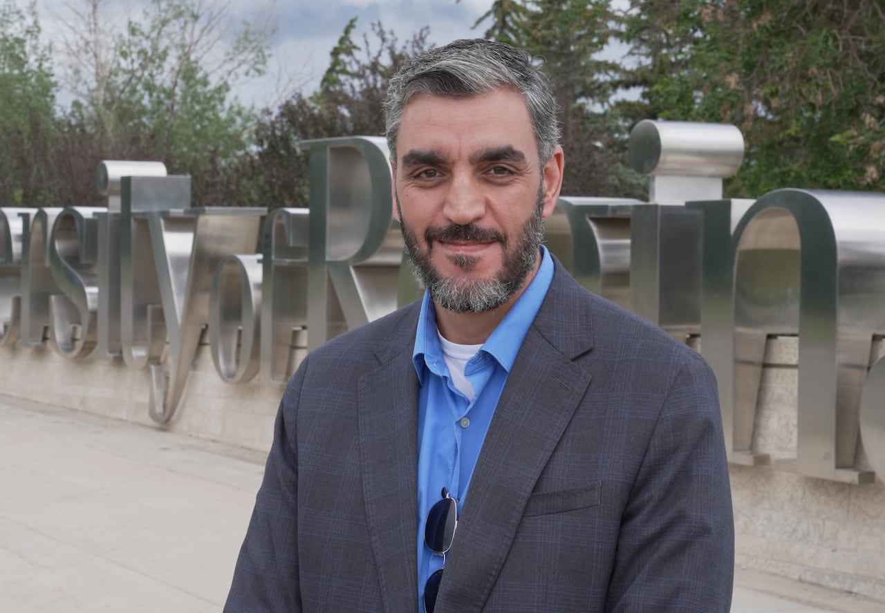 A man in a blue button-up and grey blazer smiles at the camera.