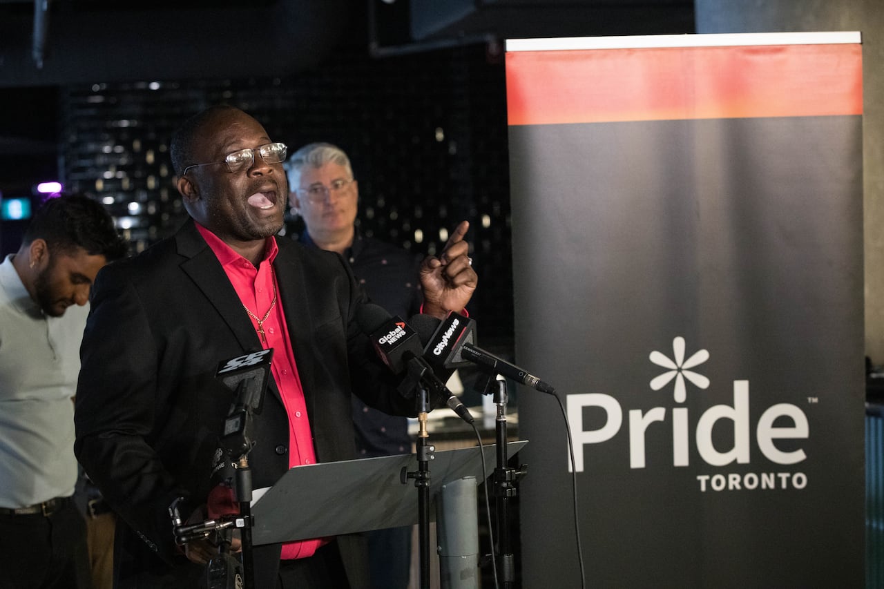 A man in a black suit and red dress shirt standing in front of a podium and sign that reads: "Pride Toronto."