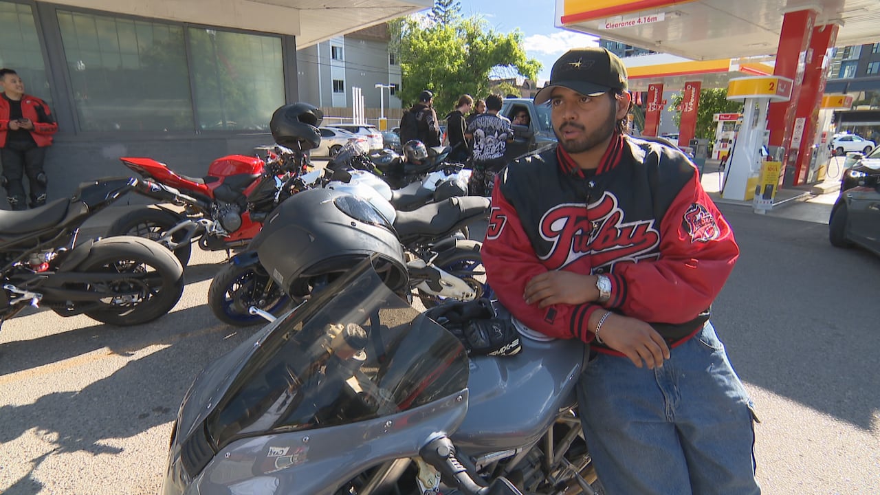 A man wearing a red and black team jacket, leaning on a motorcycle in front of a gas station.