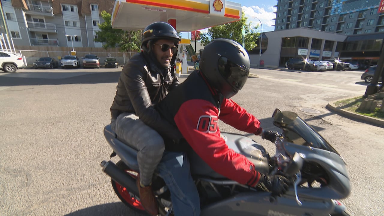 A man clings to a motorcycle driver as the pull out of a gas station.