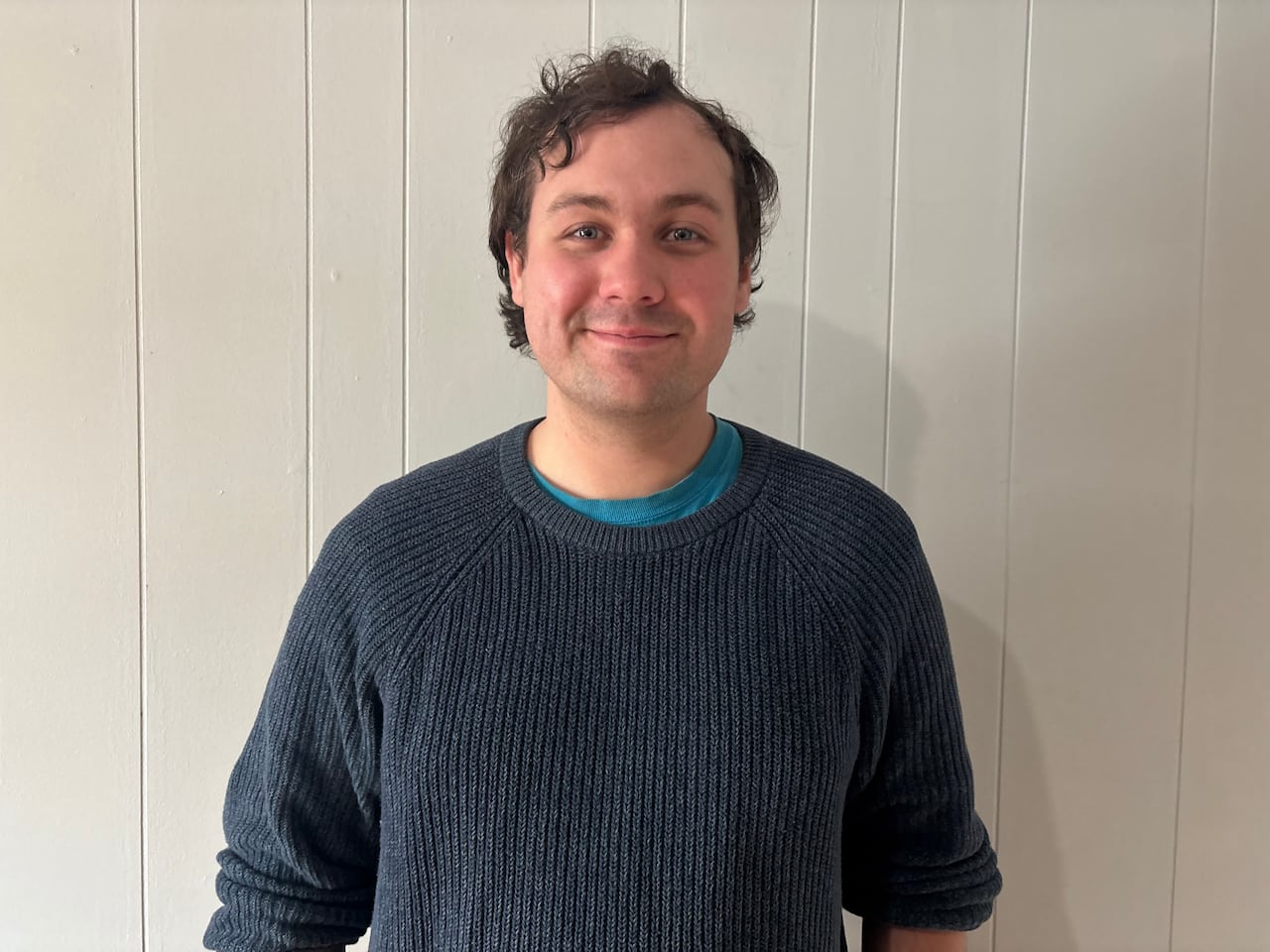 A man in a dark sweater poses in front of a white wall.