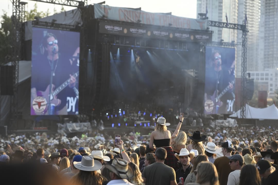 A crowd watches a concert on an outdoor stage, with many of them wearing cowboy hats.