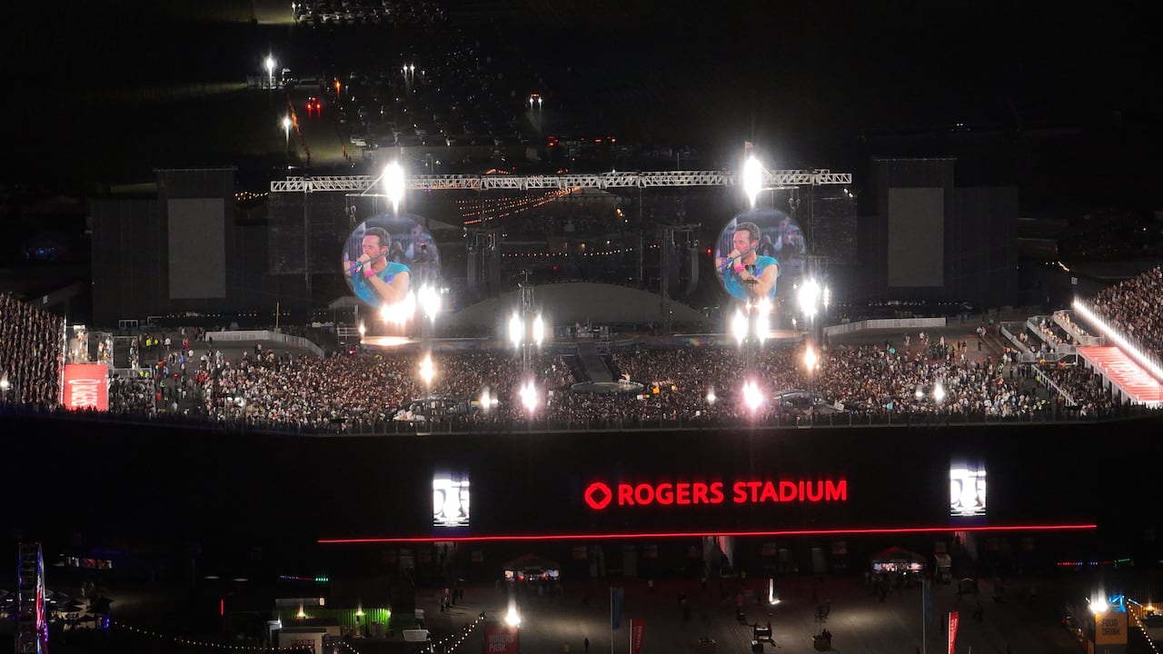 A large crowd is seen in an arena with bright lights flashing and Coldplay's Chris Martin visible on large screens.