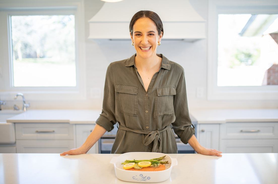 A woman in a brightly lit kitchen stands behind a counter smiling with an oven dish of fish in front of her.