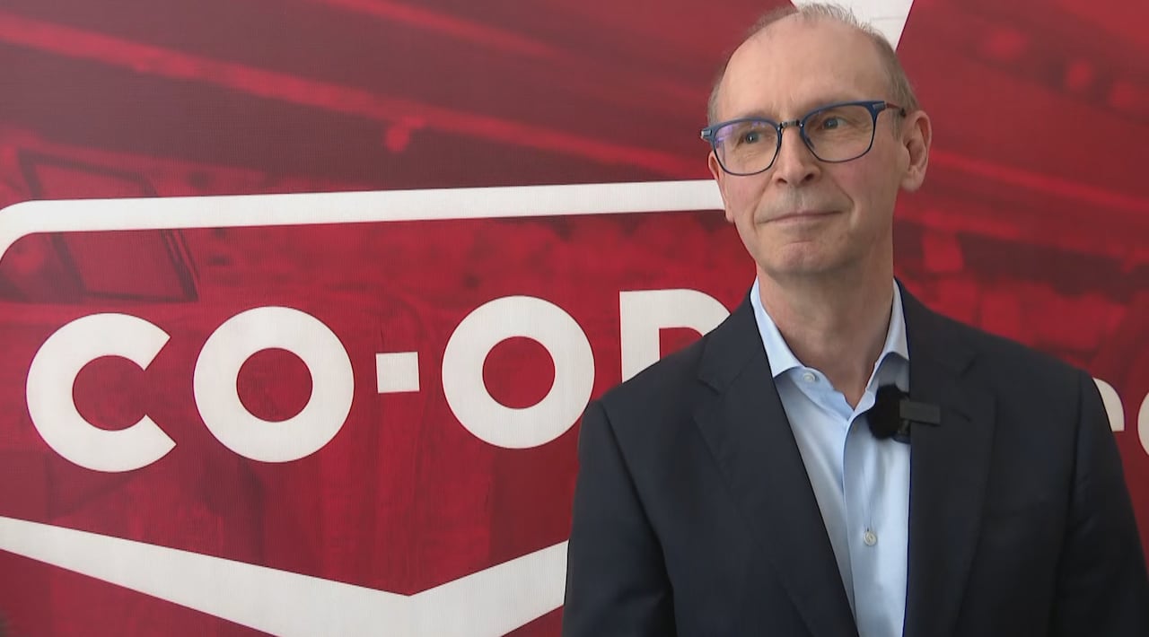 A man with glasses wears a suit and stands in front of a red banner that reads "CO-OP"