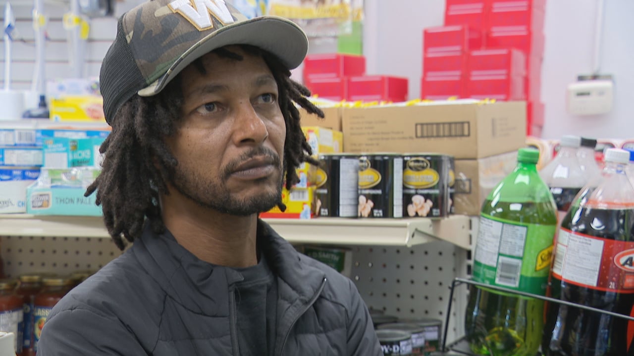A man with dark hair in short locs wears a camouflage hat with a W on it inside a convenience store
