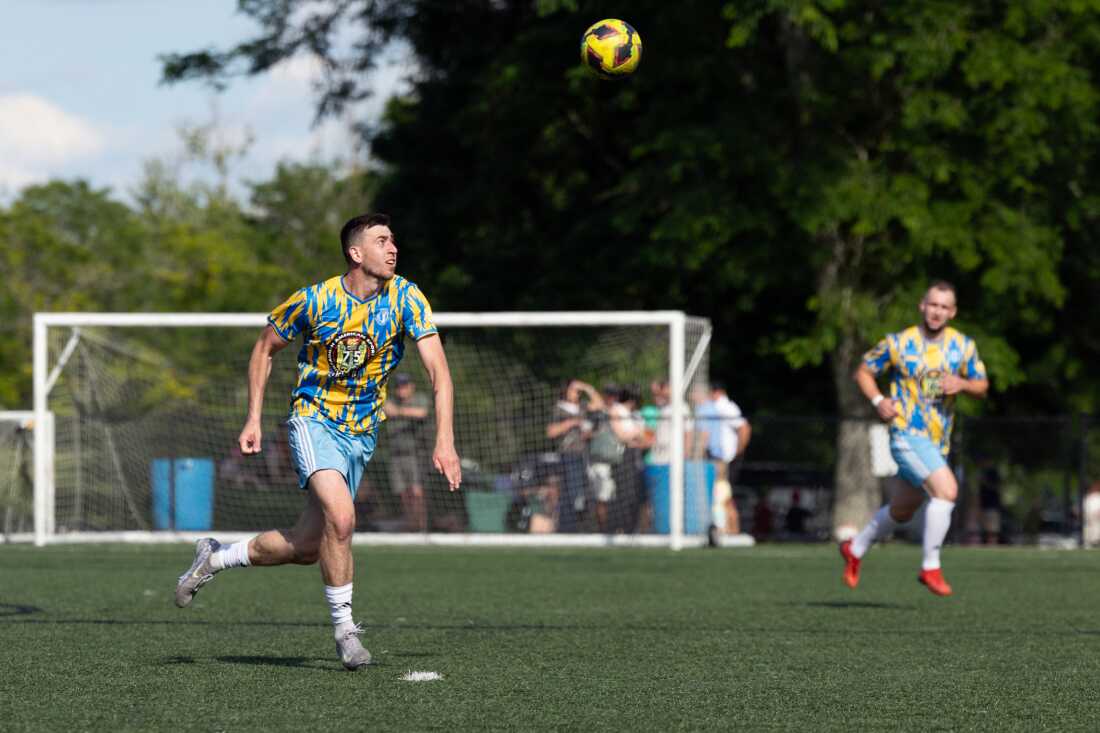 Standing on a green soccer field and wearing a yellow and light blue soccer uniform, Oleksandr “Sasha” Ostapchuk sets himself up to head a ball that's in the air while playing for a team of Ukrainian immigrants against a club with roots in Belarus at Tryzub Ukrainian American Sport Center in North Wales, Pennsylvania, on June 21.