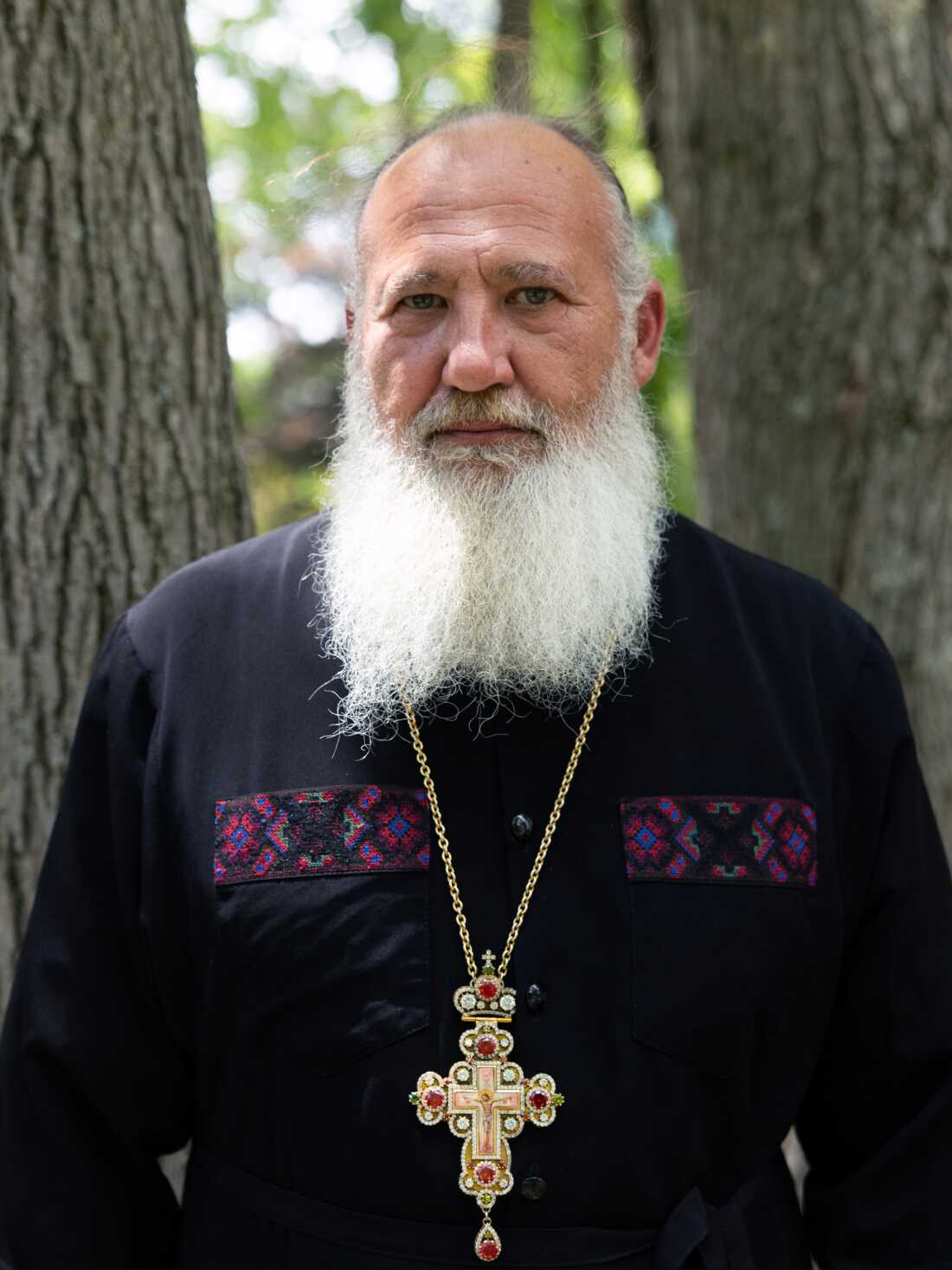 Father Taras Naumenko poses for a photo on June 21, 2026. He is wearing a cross around his neck and is photographed from about the waist up. He's standing outdoors, and two tree trunks rise behind him.