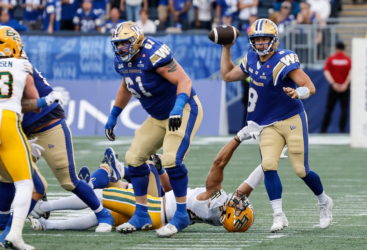 A quarterback in blue prepares to throw the ball while an opposing player, lying flat on his back, reaches up to tug on his shirt.