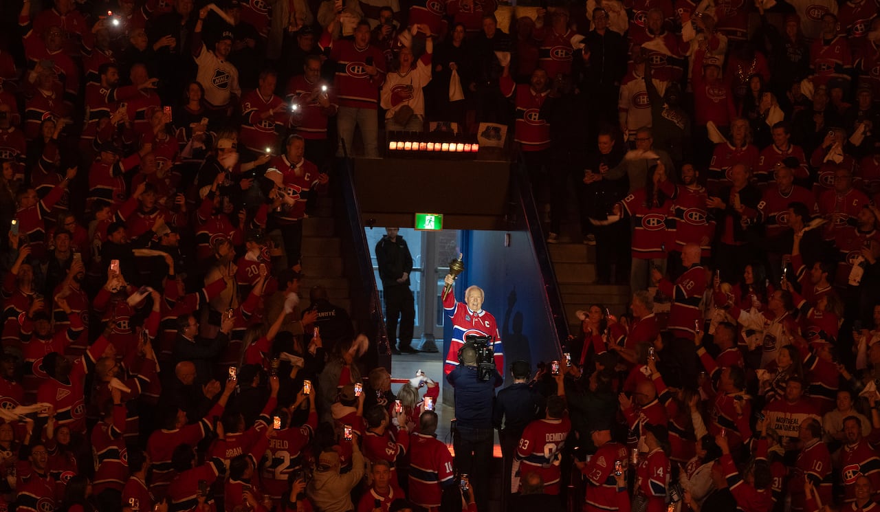 A man in a hockey sweater holds up a torch in a crowd-filled arena.