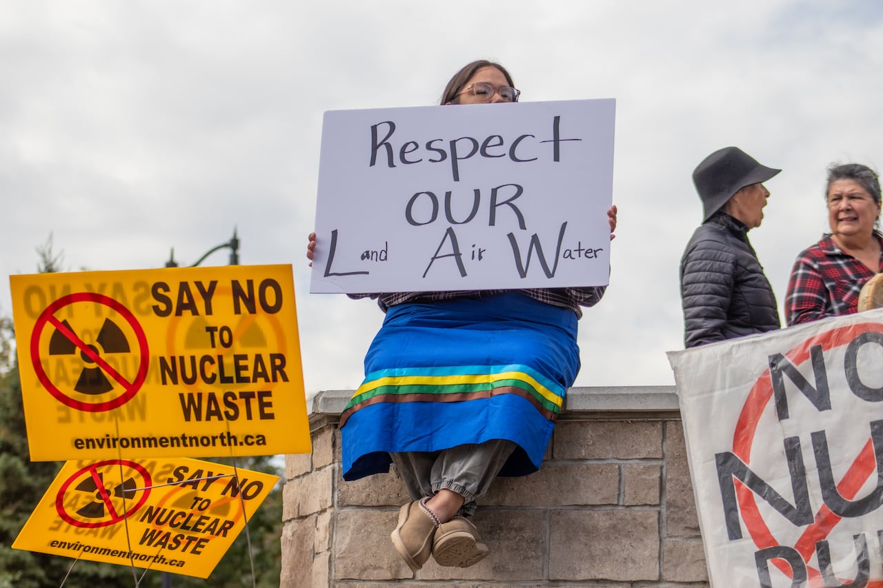 A person is seen sitting on a ledge outside, holding a sign that says 'Respect Our Law: Land, Air, Water."