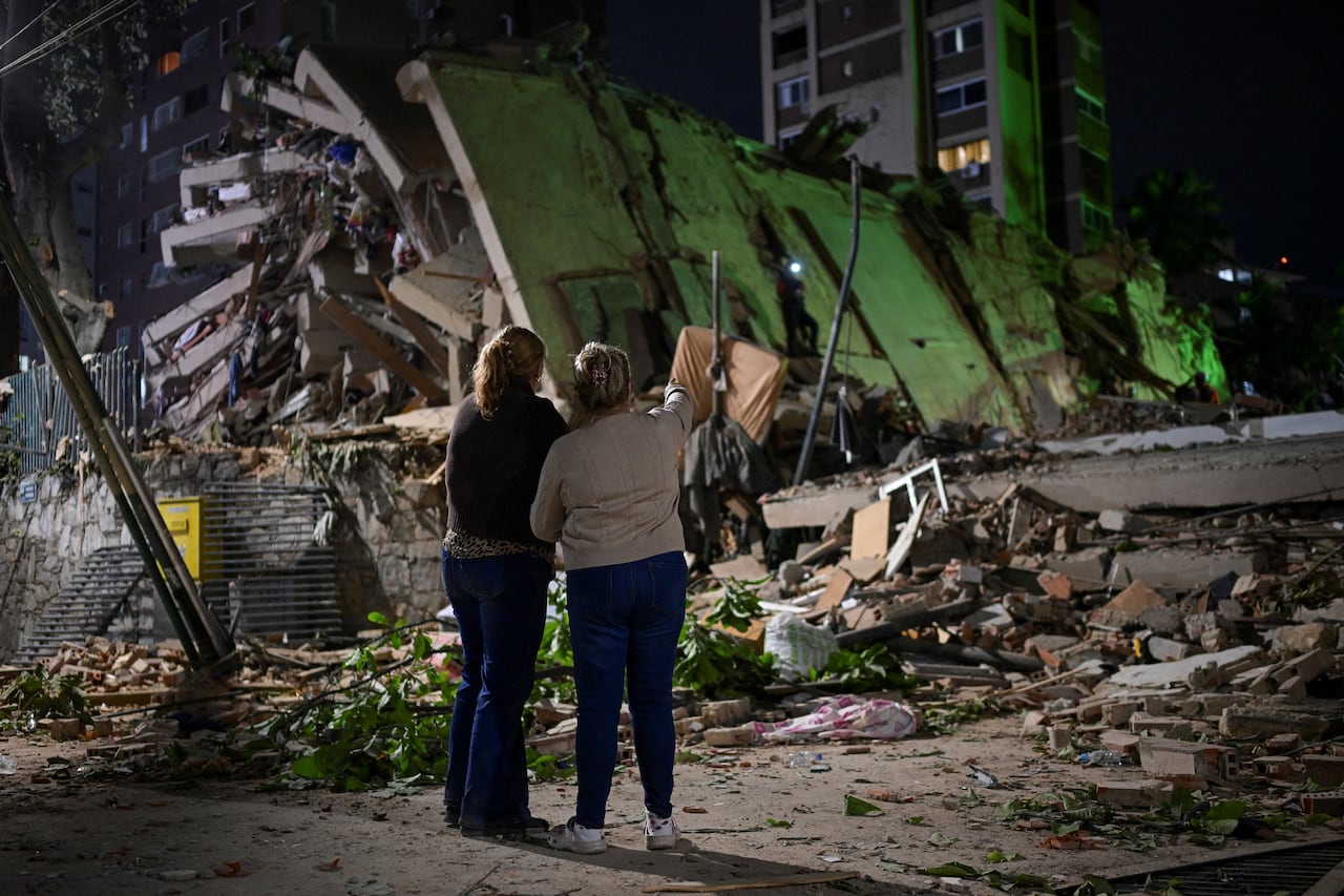 Two woman hold each other and stare at a collapsed building.