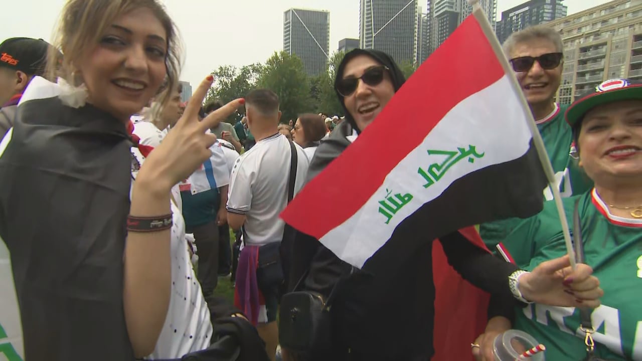 4 soccer fans in Iraq jerseys, with an Iraq flag smile at the camera, while dozens of other fans in a downtown Toronto park on an overcast summer day stand in the background.