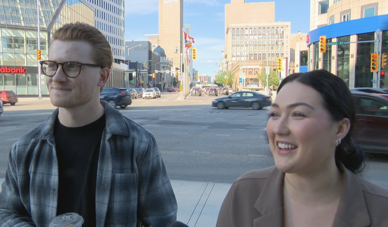 A young man and young woman stand on a street corner with traffic in the background.