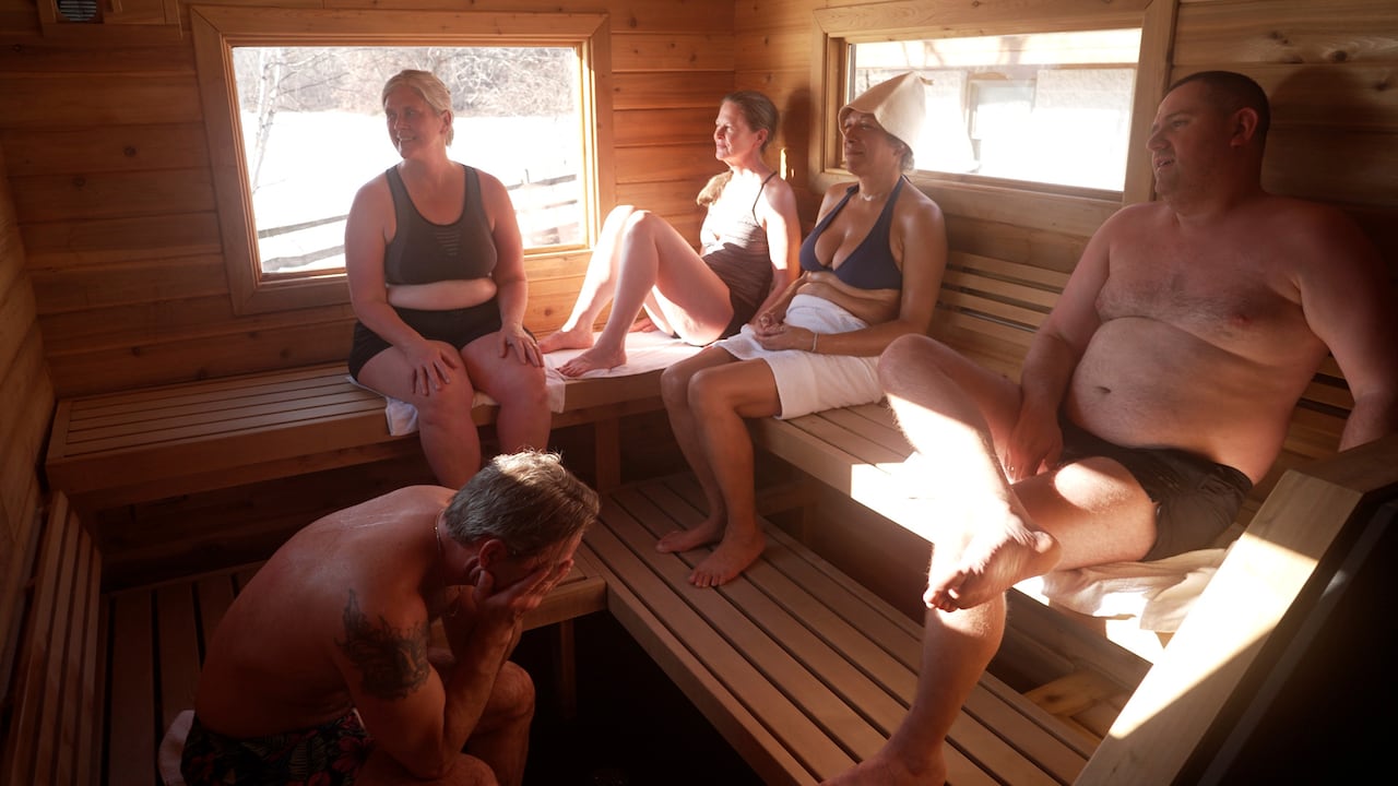 Five people sit on benches in a sauna in their bathing suits.