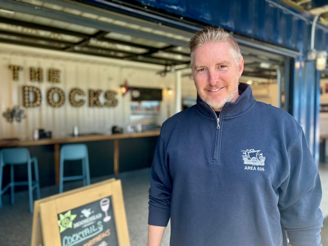 A man with blonde hair and a blue sweater smiles while standing in front of a bar.