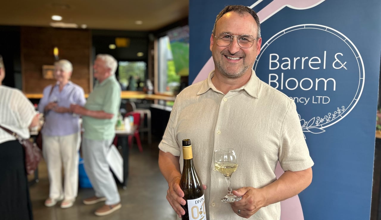 A man with a beige shirt holds a wine bottle and a wine glass in front of a sign that says "Barrel & Bloom"