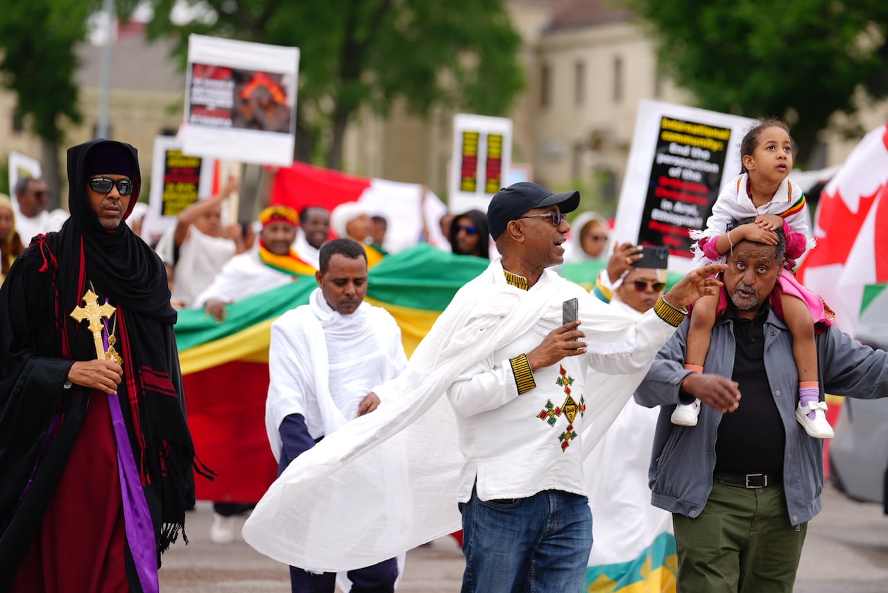 Several people marchin. The person in the middle is wearing a cape, with a shirt that has a cross on it.