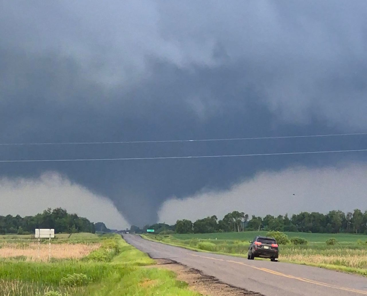 A dark blue sky and tornado are seen in the distance, looking down a highway with green fields on either side.