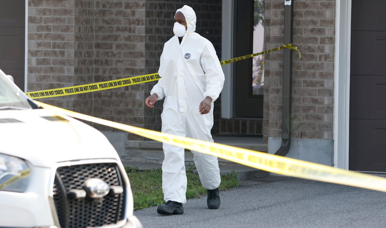 A man in a white protective suit walks behind police tape in front of a home.