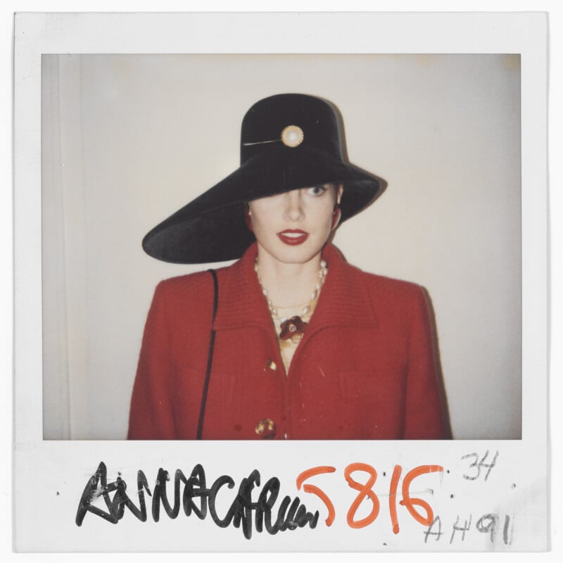 A woman in a red jacket and large black hat with a wide brim and pearl pin stands against a plain background, wearing bold red lipstick and a beaded necklace. Handwritten notes are on the photo border.