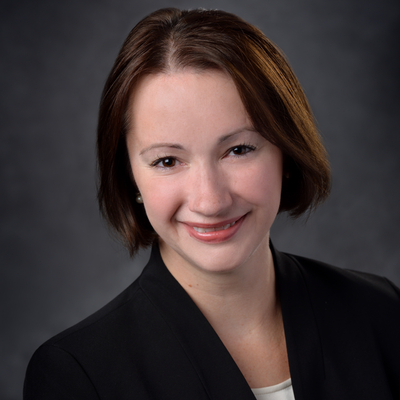 Professional headshot of a person with short brown hair wearing a dark blazer and light top against a neutral gray background.