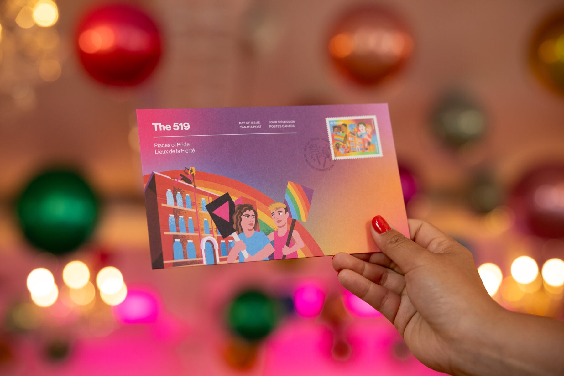 Hand holding a Pride-themed postcard with The 519 logo, celebrating LGBTQ+ community and pride events in Toronto, with colorful ornaments in the background.