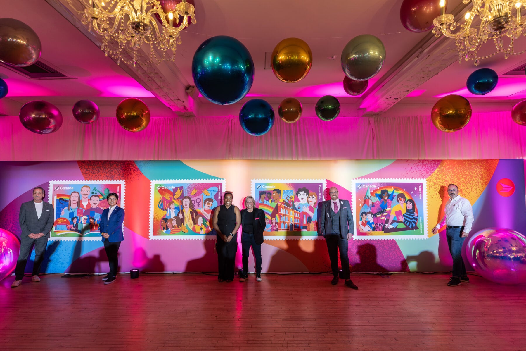 Group of people standing in front of colorful artwork at a celebration event in Toronto, with vibrant pink lighting and decorative hanging spheres, showcasing local arts and culture.