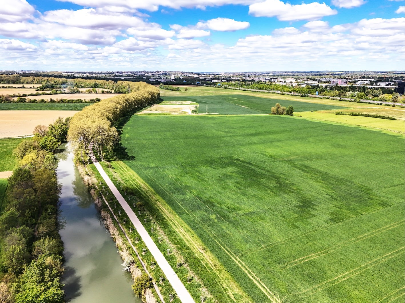 Aerial view of green fields, a tree-lined river, and a path running alongside the water under a partly cloudy sky, with distant buildings on the horizon.