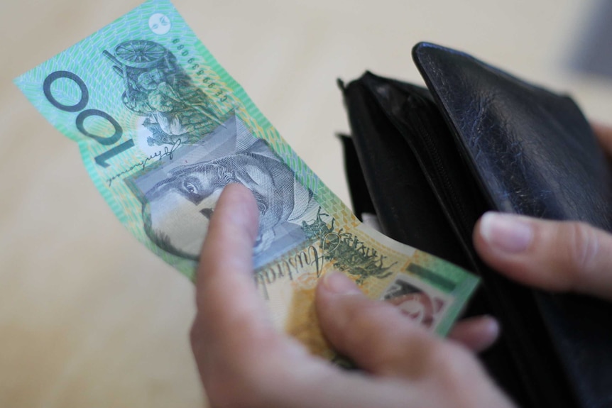 A hand holding a $100 note next to a wallet.