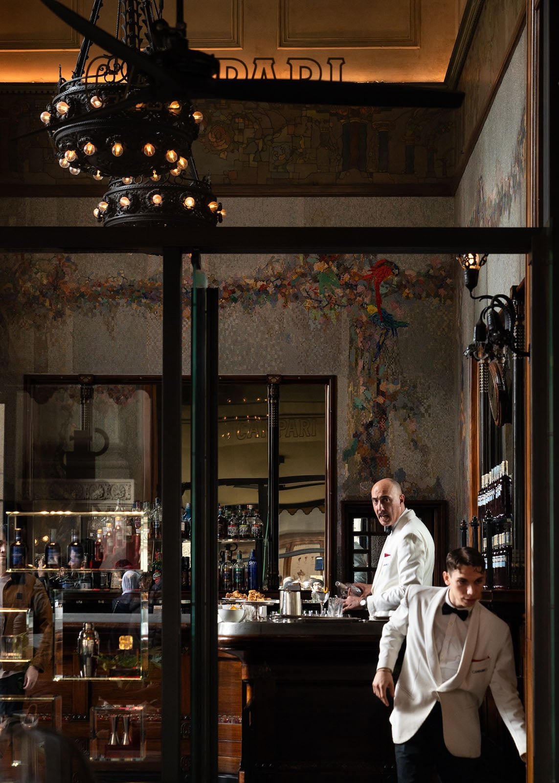 Two waiters in white jackets work behind a dark wood bar inside a vintage, elegant café with ornate lighting, wall art, and shelves of bottles. One waiter stands behind the bar, while the other walks briskly in front.
