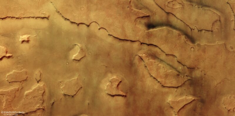 dust devils on Mars: Looking down at an orangish-brown landscape with ridges, small craters and tiny white nubs.