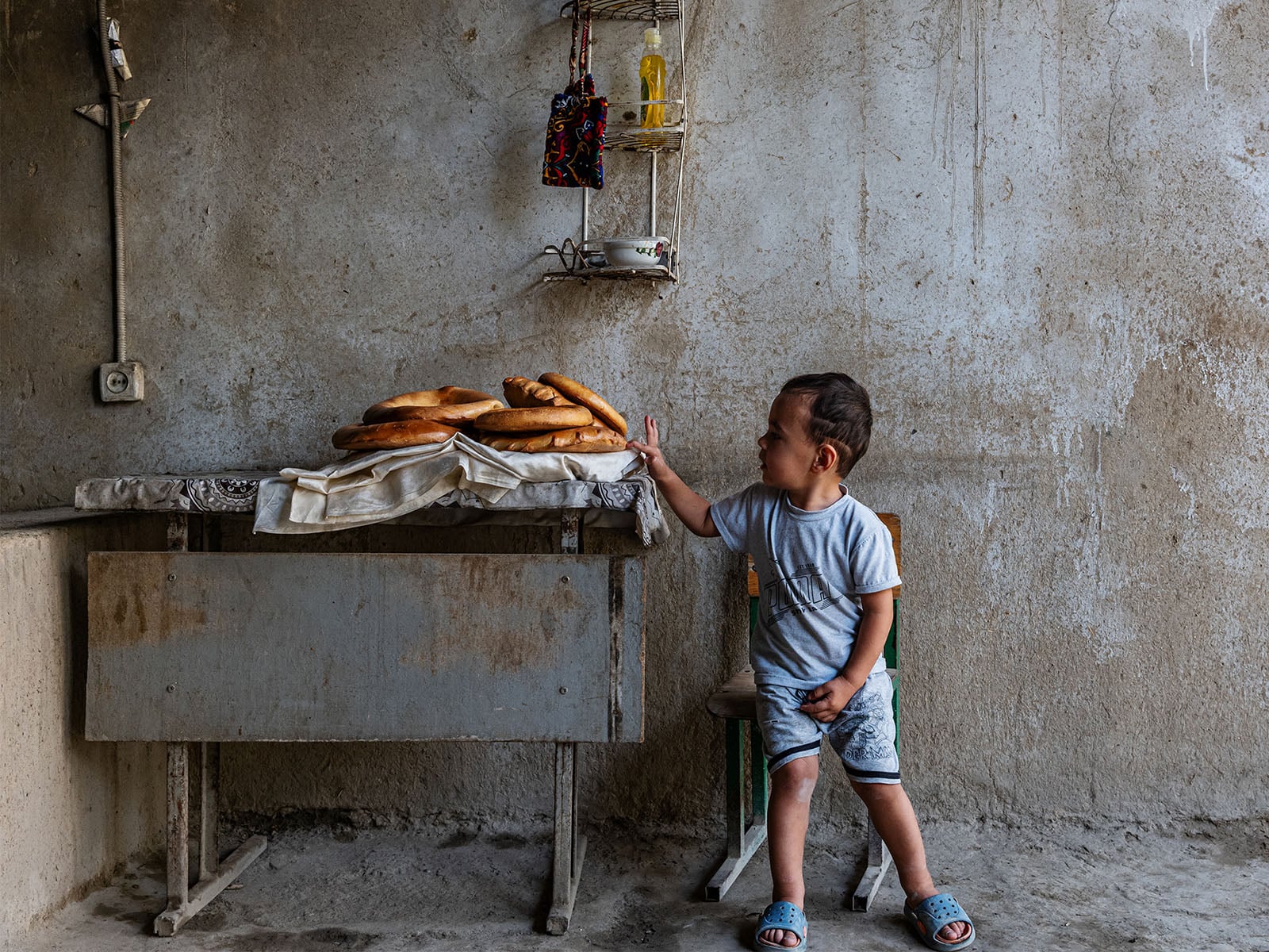 A young boy sits on a chair next to a table with several loaves of bread, reaching toward them. The scene is set against a worn, textured wall with a small shelf holding some items above the table.