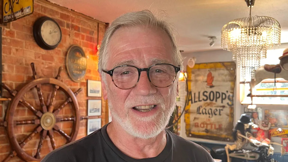 David Green, an older man with grey hair and beard and black rimmed glasses smiles at the camera, in a pub setting.