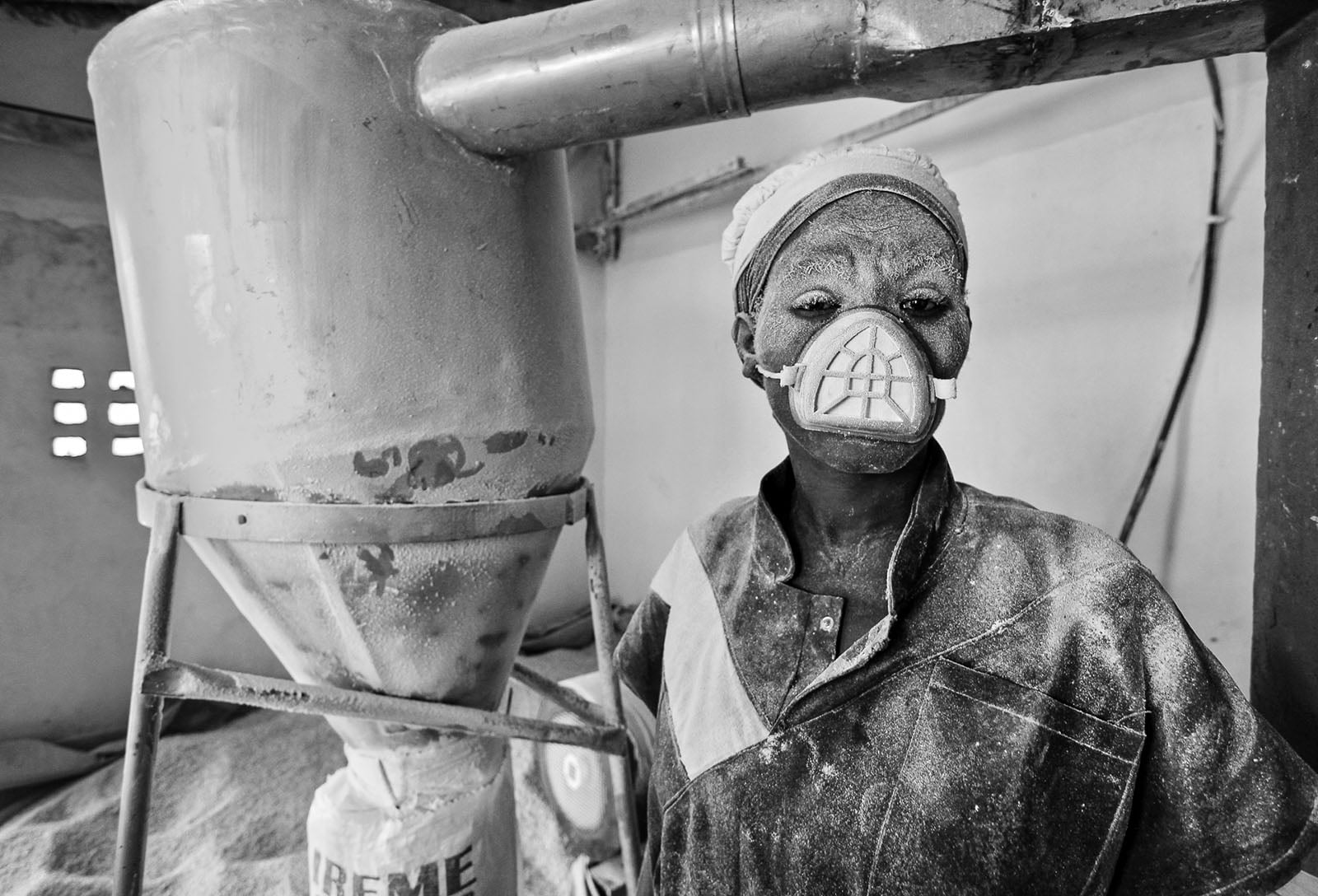 A worker covered in white powder wears a protective mask and headscarf, standing beside industrial machinery in a factory setting. The environment appears dusty, with powder on surfaces and in the air.