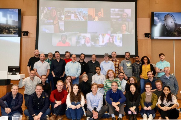 People sit in a group against a wall, a project screen with people on Zoom appears in the background