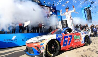 Driver stands on top of the blue‑red #57 NASCAR car during a victory celebration as confetti rains down and fans cheer behind the barriers.