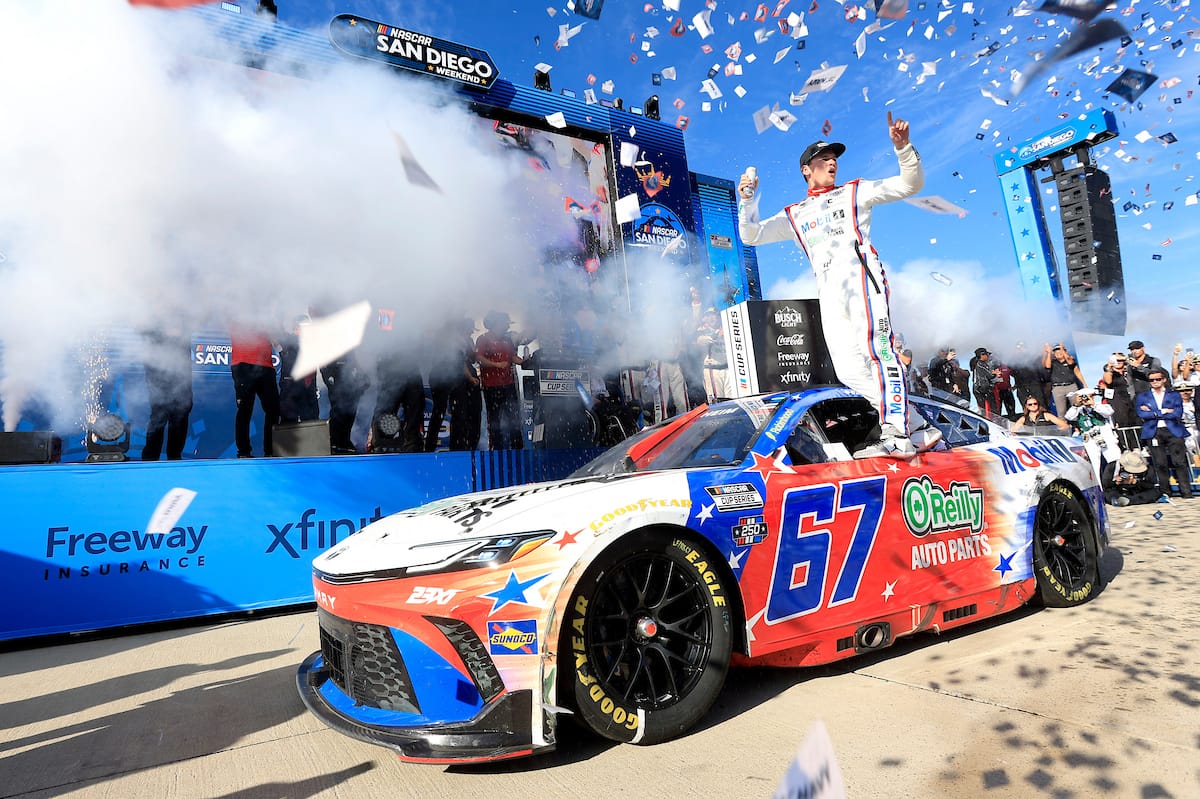 Driver stands on top of the blue‑red #57 NASCAR car during a victory celebration as confetti rains down and fans cheer behind the barriers.