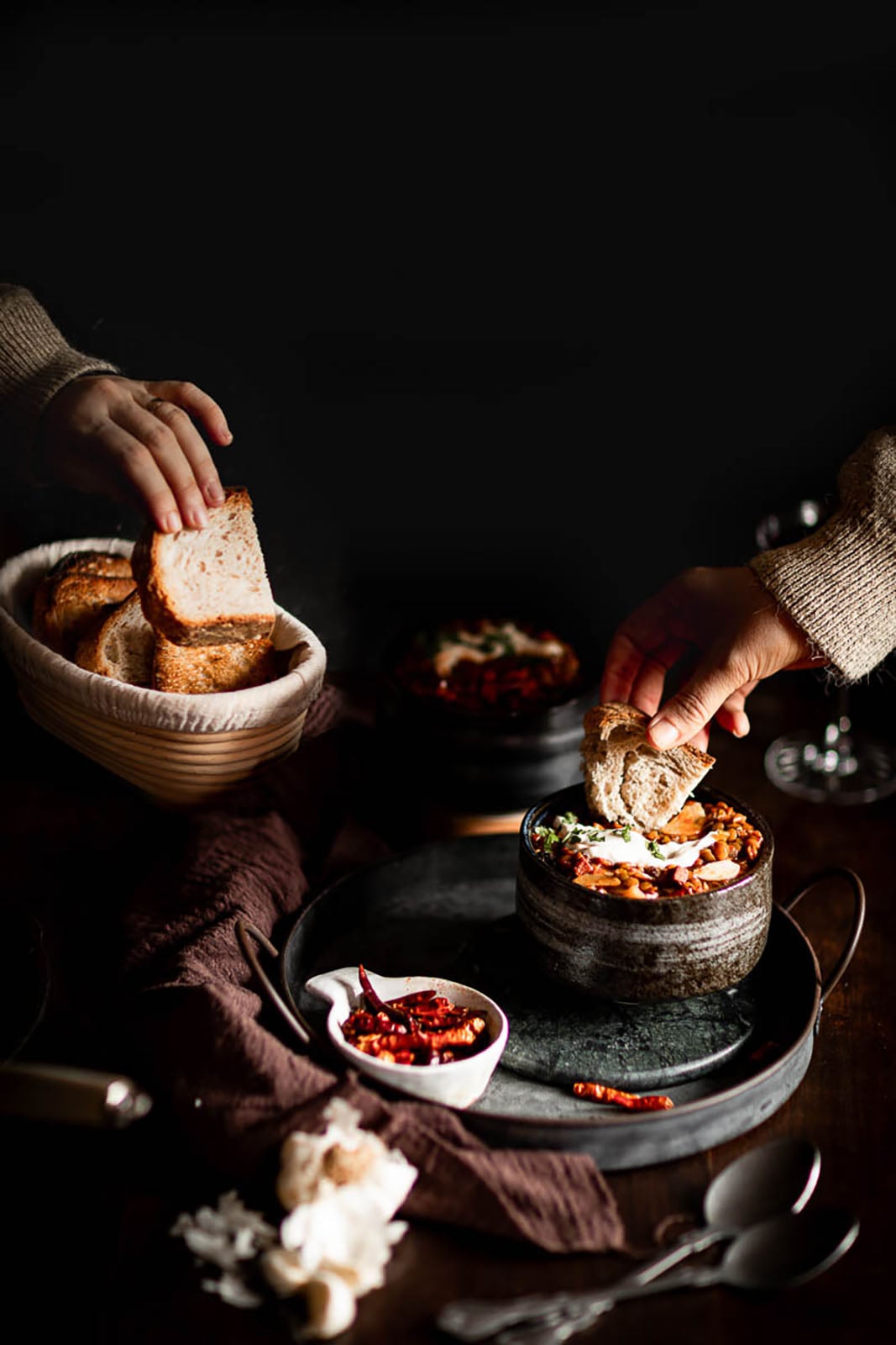 Two hands reaching for rustic bread; one takes slices from a basket, the other dips bread into a bowl of stew on a dark table with small dishes of dried chili and a brown cloth.