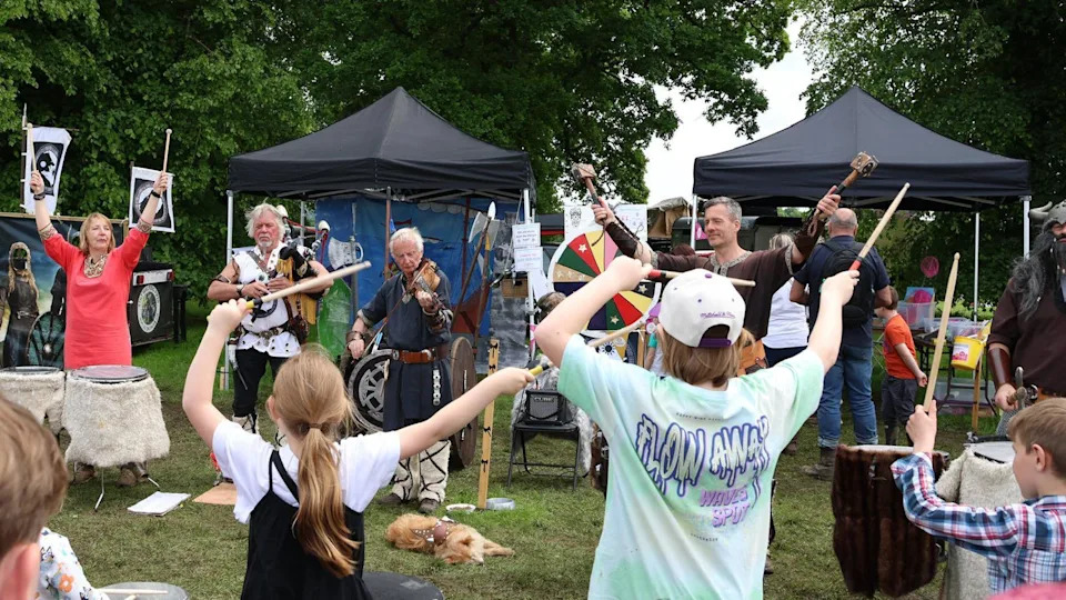 In a field, the Biking Vikings, dressed as Vikings, are performing in a line playing various instruments including the pipes, drums and strings. There are children facing them, holding drumsticks, with their arms raised too. 