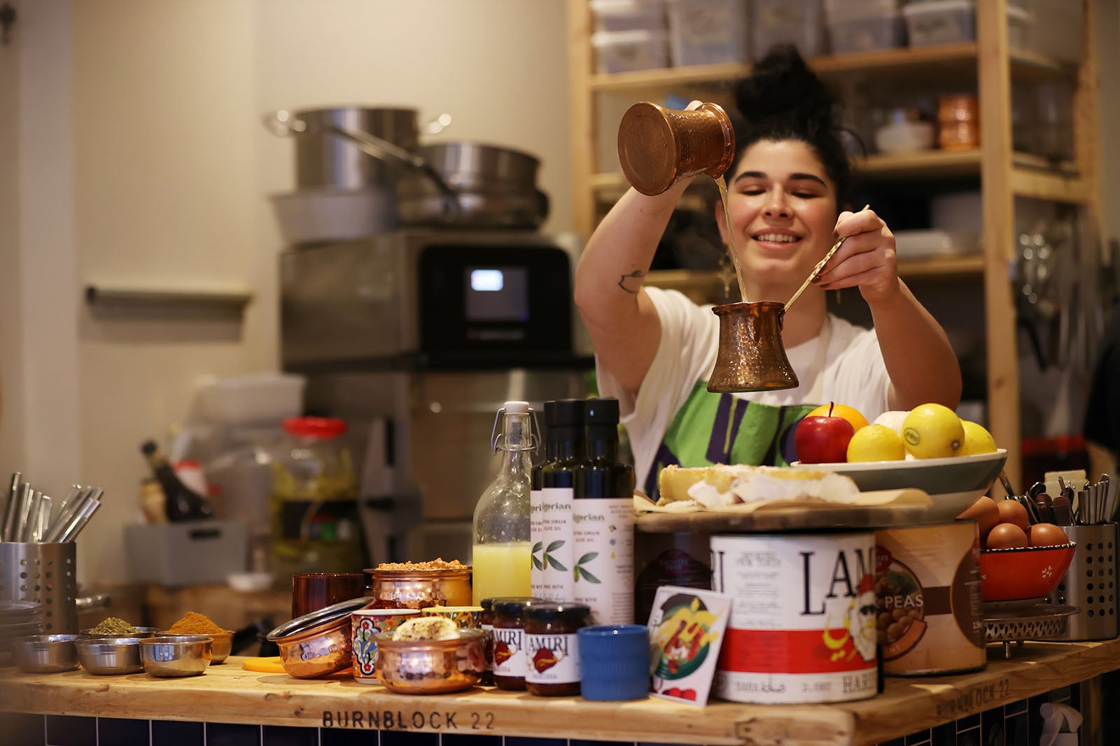 A smiling woman in a kitchen pours coffee from a long-handled copper pot. The counter in front of her is filled with various foods, canned goods, fresh fruit, eggs, and bottles. Shelves with kitchenware are in the background.