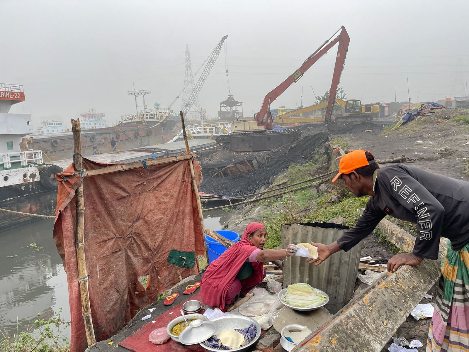 A woman serves food from a roadside stall beside a river, handing a plate to a man. Industrial cranes, ships, and coal are visible in the misty background, indicating a busy worksite near the water.