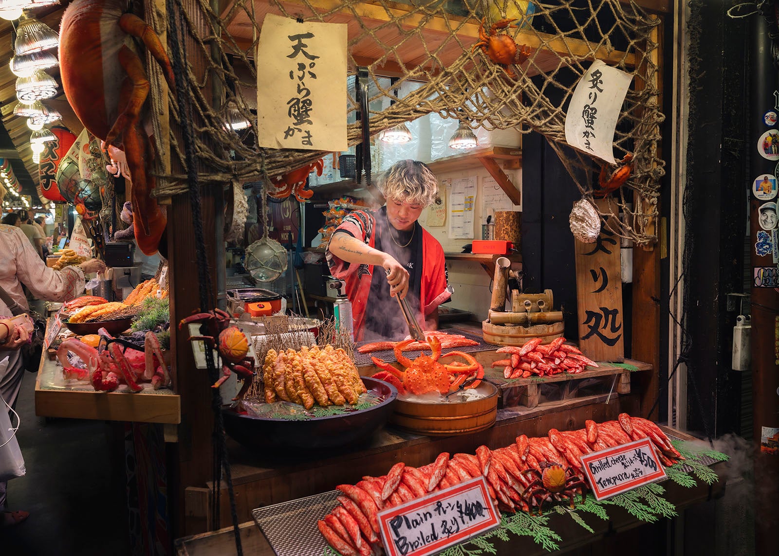A street food vendor cooks seafood at a colorful market stall in Japan, surrounded by displays of crab, shellfish, and food signs written in Japanese. The scene is lively and full of vibrant colors and textures.