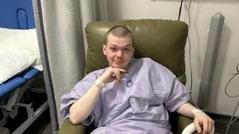 A man wearing a purple hospital gown sits next to a bed