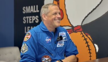 A man in a blue jumpsuit sits smiling in front of a wall with a giant snoopy astronaut painted on.